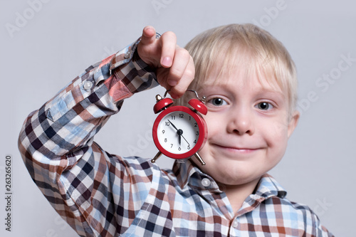 Little blond boy with a red alarm clock in his hands. Morning concept. Light background