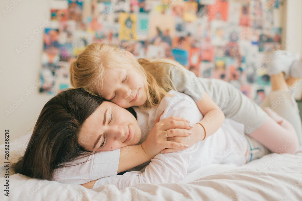 Lifestyle soft focus portrait of happy mom hugs her adorable young daughter on white bed. Morning family portrait of smiling mother and her funny daughter hugs on 90s style wall on background. Lifestyle soft focus portrait of happy mom hugs her adorable young daughter on white bed. Morning family portrait of smiling mother and her funny daughter hugs on 90s style wall on background.