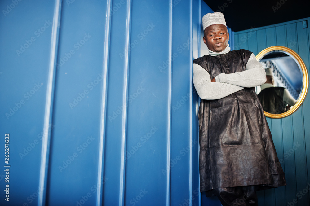 African men in black traditional clothes with cap against mirror on blue wall.