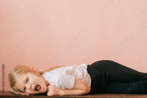 Lifestyle portrait of clowning little girl make faces and shouting on pink wall background. Funny expressive female with ridiculous face kid lying on table and screaming like crazy with gesturing hand