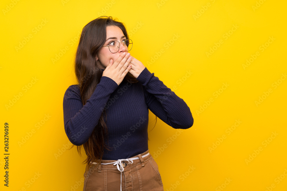 Teenager girl over isolated yellow wall covering mouth and looking to the side Teenager girl over isolated yellow wall covering mouth and looking to the side