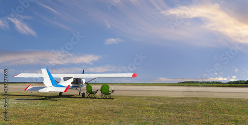 Rear side view of light aircraft with piston engine on airfield with two folding lounge chairs under the wing against the backdrop of a calm cloudy sky