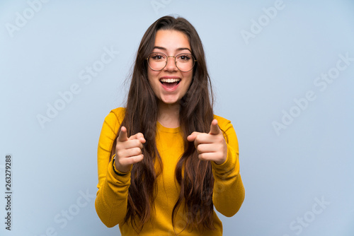 Teenager girl over isolated blue wall points finger at you