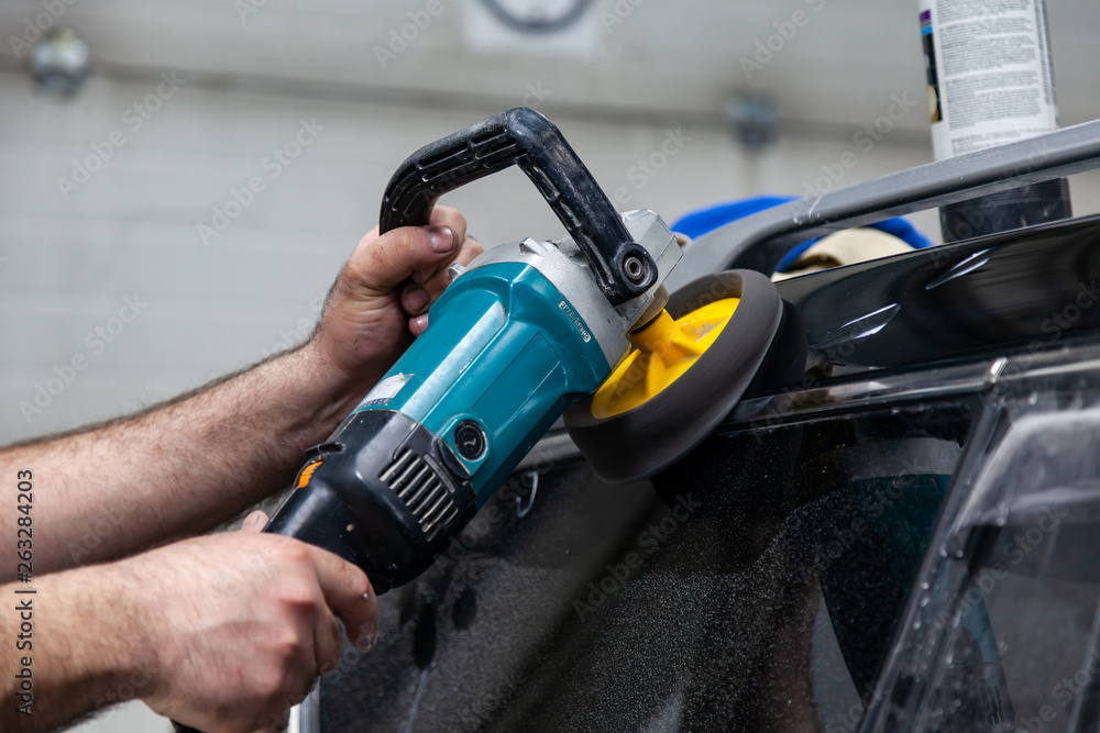 Close-up view on the hands of a man worker who holds a tool polisher for polishing the roof of a car while working in a vehicle detailing workshop. Auto service industry.