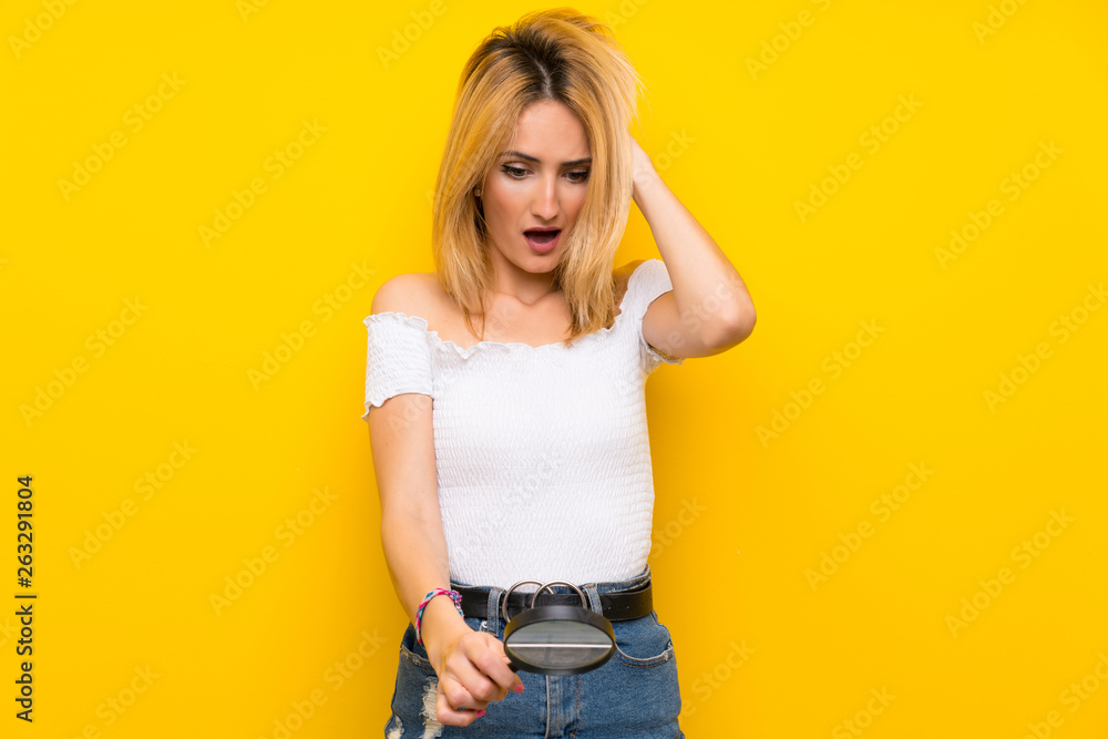 Young blonde woman over isolated yellow wall holding a magnifying glass Young blonde woman over isolated yellow wall holding a magnifying glass