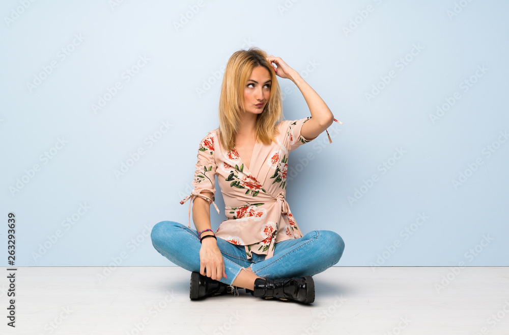 Young blonde woman sitting on the floor having doubts while scratching head Young blonde woman sitting on the floor having doubts while scratching head