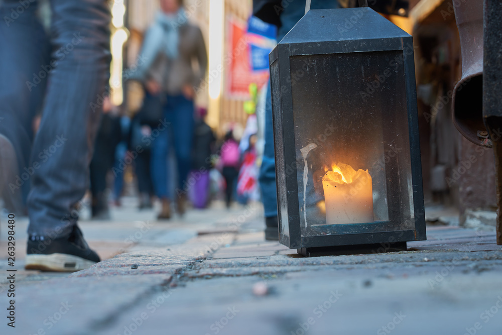 Obraz premium Big metal dirty lantern with burning candle inside, outdoor on a ground with blurred crowded street on a background.
