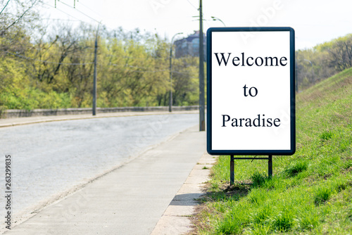 Vertical billboard with an inscription on a white background "WELCOME TO PARADISE", stands on the side of the road from the pavement