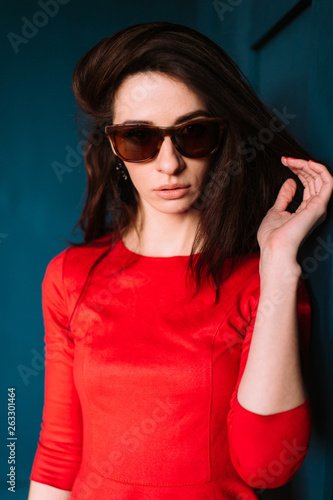 Beautiful fashion girl with dark long hair, spanish appearance in sunglasses and red elegant dress posing on blue wall in studio. Indoor lifestyle portrait of stylish woman. Aamzing brunette model
