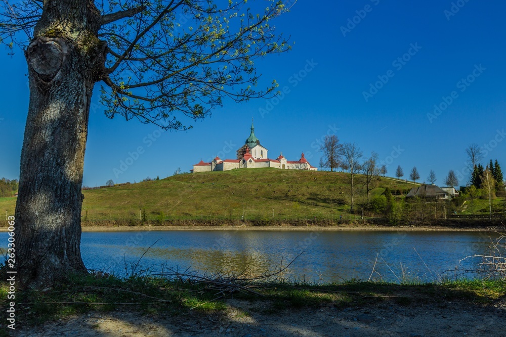 Naklejka premium Spring view of Pilgrimage Church of St John of Nepomuk at Zelena Hora on top of a green hill at Zdar nad Sazavou, bright sunny day with clear blue sky, Convent lake, tree and sandy land in foreground