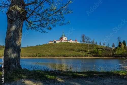 Spring view of Pilgrimage Church of St John of Nepomuk at Zelena Hora on top of a green hill at Zdar nad Sazavou, bright sunny day with clear blue sky, Convent lake, tree and sandy land in foreground