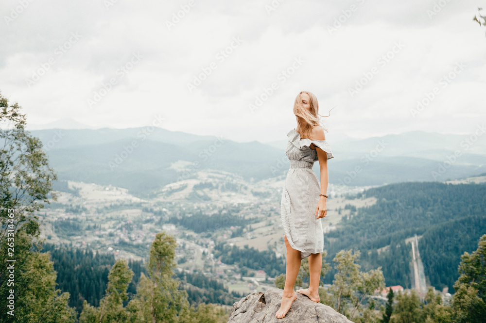 Naklejka premium Young beautiful barefoot blonde girl with long hair in summer dress standing on top of conquered mountain at stone and enjoying fabulous landscape scenic view with mountains and village in valley