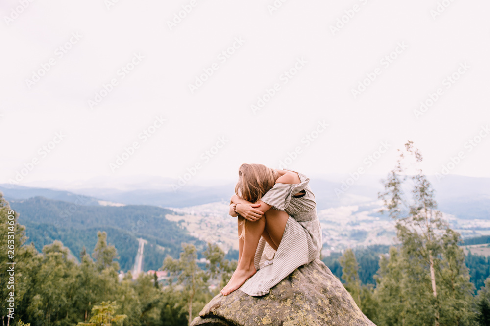 Young girl sits at stone on top of mountain and hugs her legs with head lying on knees. Beautiful blonde woman in depression hiding her face. Psychological female outdoor portrait. Loneliness concept.
