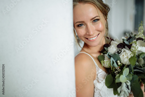 Magnificent bride with beautiful make-up and cute smile holding wedding bouquet in her hands and looking into camera