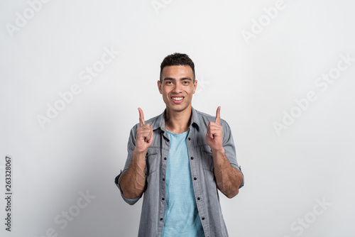 Fotografie Pretty young man  showes gesture with index fingers  points upwards over white b