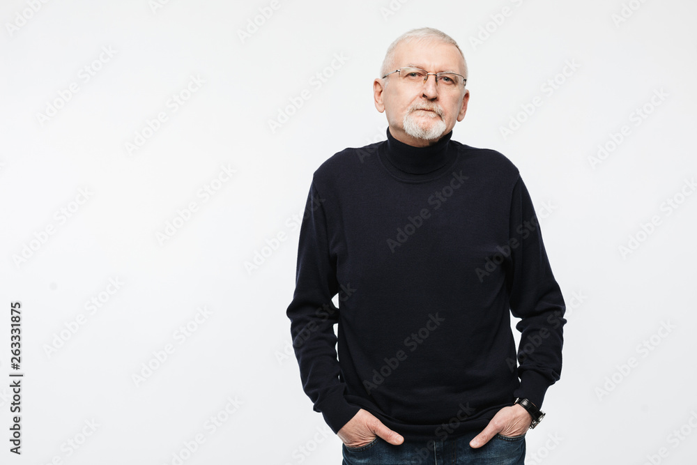 Old attractive man with gray hair and beard in eyeglasses and black sweater thoughtfully looking in camera while holding hands in pockets over white background Old attractive man with gray hair and beard in eyeglasses and black sweater thoughtfully looking in camera while holding hands in pockets over white background