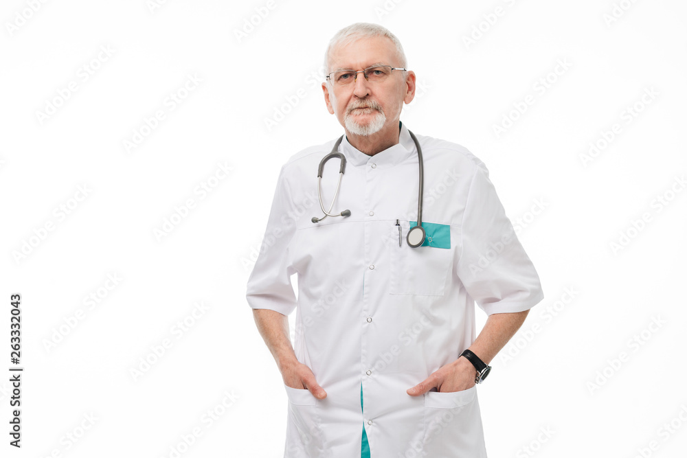 Old handsome doctor with gray hair and beard in eyeglasses and uniform holding hands in pockets while dreamily looking in camera over white background Old handsome doctor with gray hair and beard in eyeglasses and uniform holding hands in pockets while dreamily looking in camera over white background