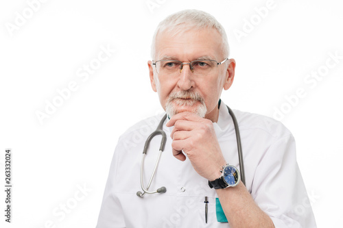 Old handsome doctor with gray hair and beard in eyeglasses and uniform holding hand near chin while thoughtfully looking aside over white background