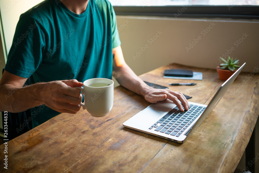 Trendy Caucasian male working on laptop and drinking tea from home on a wooden table Trendy Caucasian male working on laptop and drinking tea from home on a wooden table