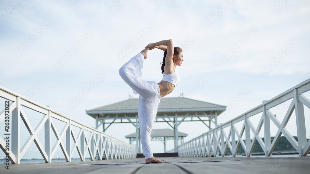 Young woman in white yoga clothes is standing in asana Natarajasana exercise on wooden floor near sea. on a pier on Maldives. Amazing yoga landscape and enjoying sea view