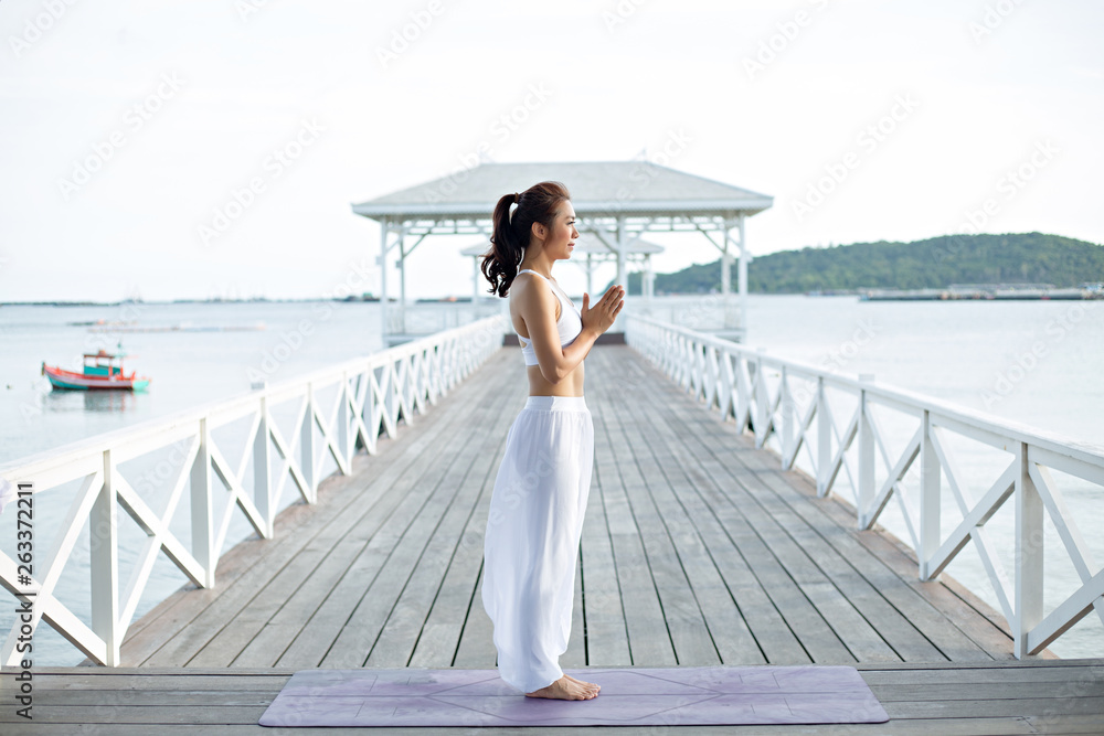 Young woman in white yoga clothes is staying and doing meditation on the beach near sea. on a pier on Maldives. Amazing yoga landscape and enjoying sea view, concept for exercising, health care