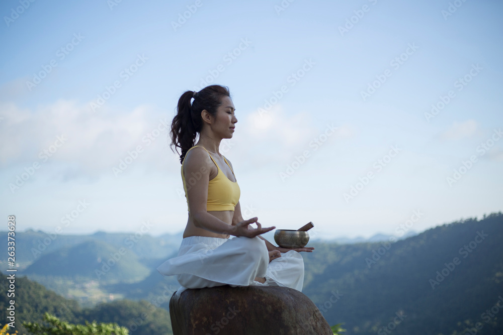 Young woman doing exercise yoga. sitting on the rock with tibetan singing bowl with its mallet singing bowl,Lotus pose. (The Nepal word on the tibetan singing bowls means wish everything is fine.)