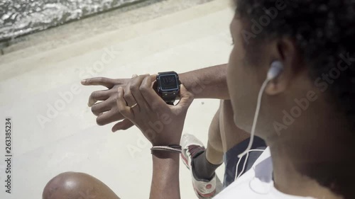 Focused African American man setting sport watch. Young guy sitting on embankment and using modern device. Technology concept photo