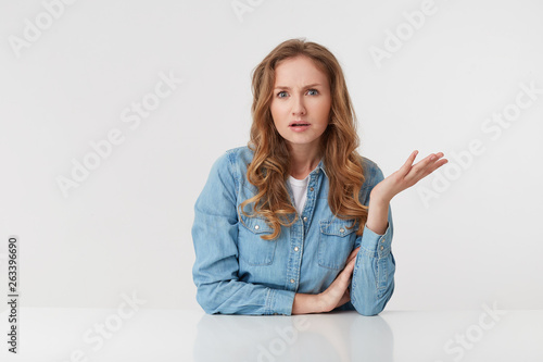 Portrait of confused young woman with long blond wavy hair, sitting at the table, one palm raised, looks skeptically displeased indignantly with incomprehension, over white background.