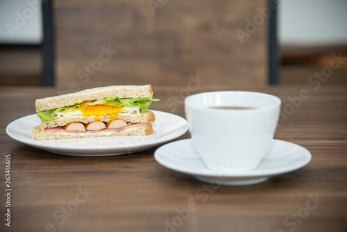 Sandwich and cup of coffee on white plate and wooden table, sandwich is made from bread, sausage, fried egg and vetgetable is American breakfast or fast food is unhealthy food or junk food