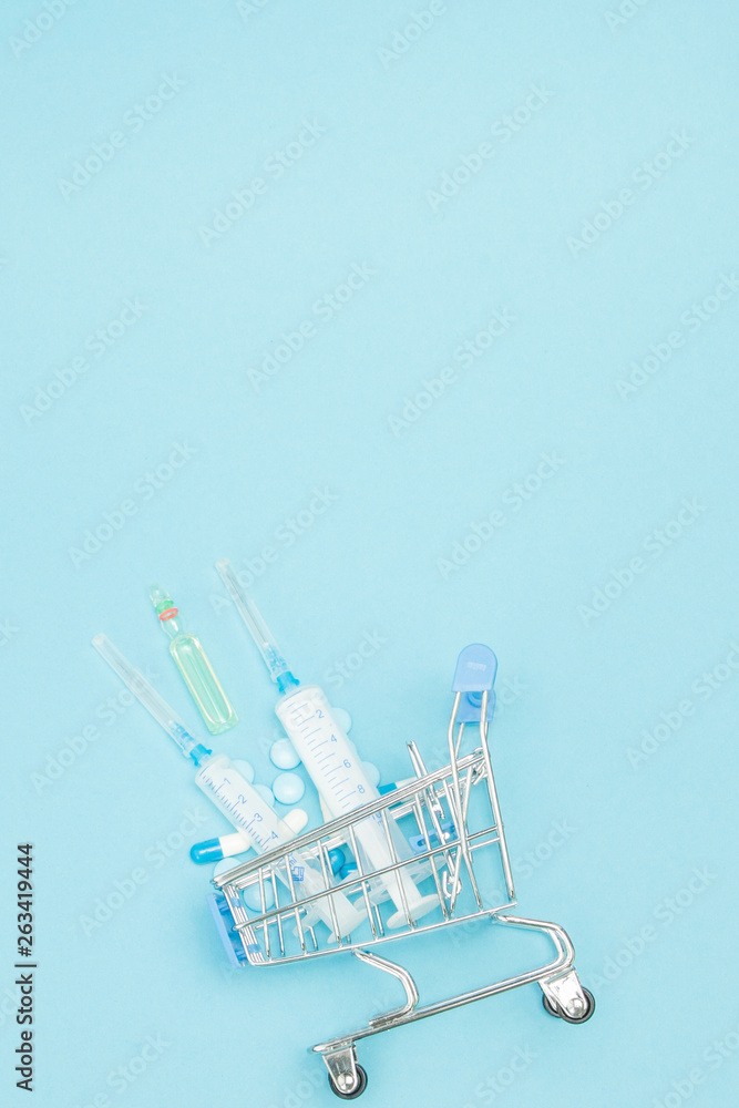 Pills and medical injection in shopping trolley on blue background. Creative idea for health care cost, drugstore, health insurance and pharmaceutical company business concept. Copy space Pills and medical injection in shopping trolley on blue background. Creative idea for health care cost, drugstore, health insurance and pharmaceutical company business concept. Copy space