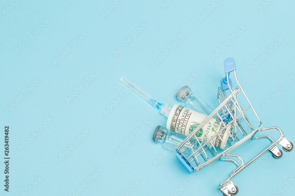 Obraz premium Medical injection and dollars in shopping trolley on blue background. Creative idea for health care cost, drugstore, health insurance and pharmaceutical company business concept. Copy space