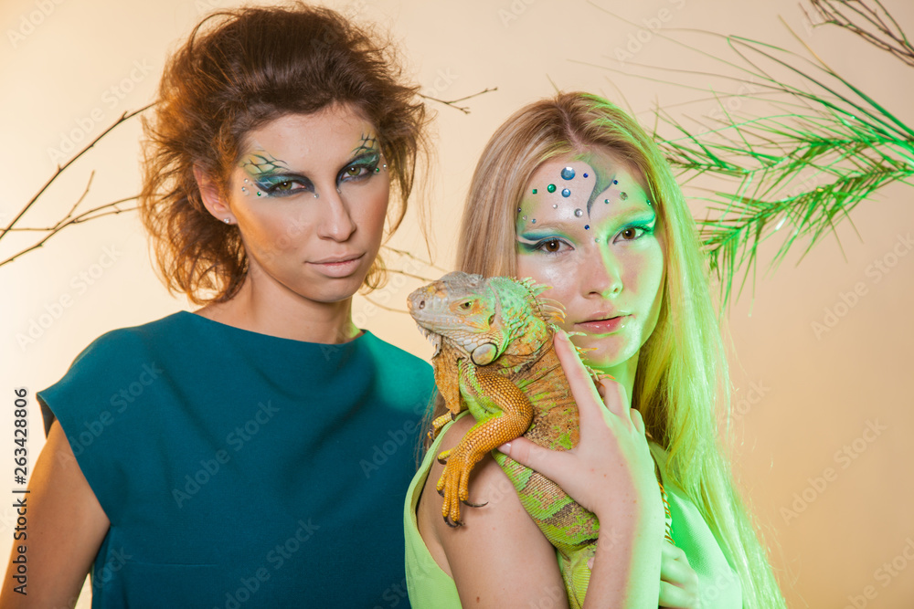 Wild animal in the hands of man. Two girls with an unusual make-up and with an iguana in their hands. Animal out of it. An image of a female predator and snake Wild animal in the hands of man. Two girls with an unusual make-up and with an iguana in their hands. Animal out of it. An image of a female predator and snake