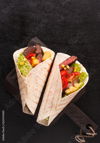 Mexican tacos with beef, vegetables and pineapples and corn grains on a wooden kitchen Board. Taco al pastor on a wooden Board on a black background. Top view with copy space
