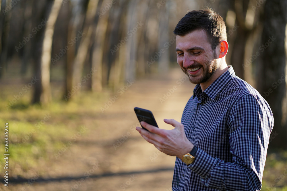 Attractive bearded caucasian man with freckles with short brown hair and green eyes in plaid shirt with golden watch reads a message on the phone and laughs in the autumn forest in the rays of sun. Attractive bearded caucasian man with freckles with short brown hair and green eyes in plaid shirt with golden watch reads a message on the phone and laughs in the autumn forest in the rays of sun.