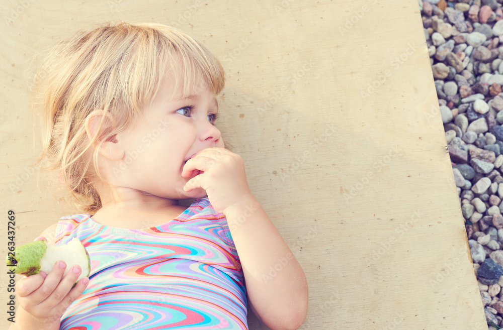 Cute adorable toddler girl eating fresh pear lying on the beach. Cute adorable toddler girl eating fresh pear lying on the beach.