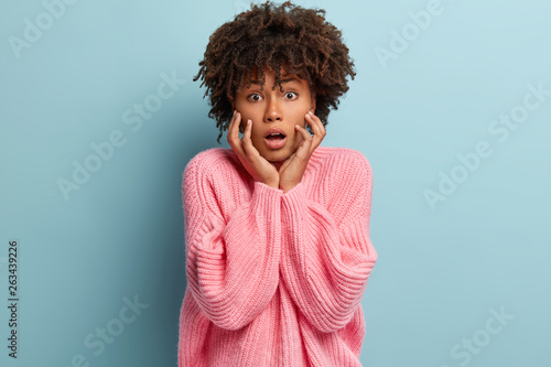 Indoor photo of surprised emotional dark skinned woman keeps mouth opened  touches cheeks  expresses great disbelief  astonished by latest news  isolated over blue background. Facial expressions