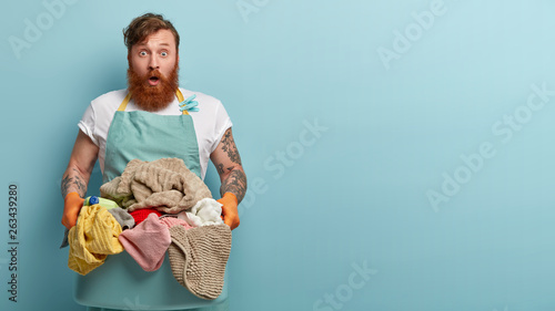 Stupefied foxy housekeeper keeps jaw dropped, holds basin with dirty laundry, has cleaning detergent, wears casual domestic clothes, isolated over blue background with free space aside for text