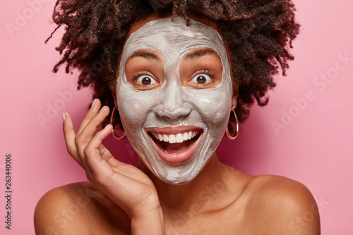 Close up portrait of delighted Afro American lady has white clay mask on face, smiles broadly, surprised to have fresh skin after beauty procedures, has consultancy with beautician or cosmetologist