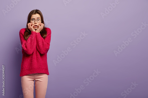 Horizontal shot of nervous stressed woman feels neurotic, bites finger nails, wears oversized long red jumper, reacts on astonishing news, stands over purple background. Empty space for information photo