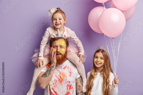 Three family members pose indoor over purple background. Funny two daughters and dad have fun, color faces with colorful watercolors, celebrate Childrens Day together. Entertainment concept.
