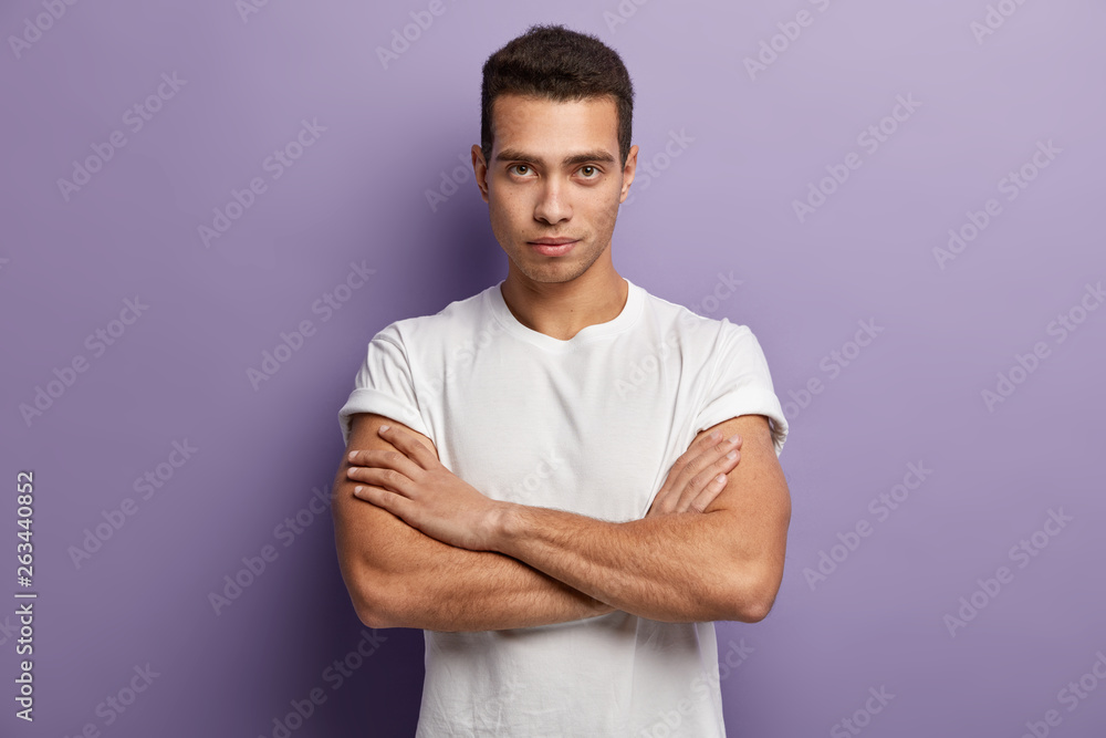 Self assertive serious young guy keeps muscular arms crossed over chest, looks directly at camera, gazes attentively something, dressed in casual white t shirt, looks confident. I am ready listen you Self assertive serious young guy keeps muscular arms crossed over chest, looks directly at camera, gazes attentively something, dressed in casual white t shirt, looks confident. I am ready listen you