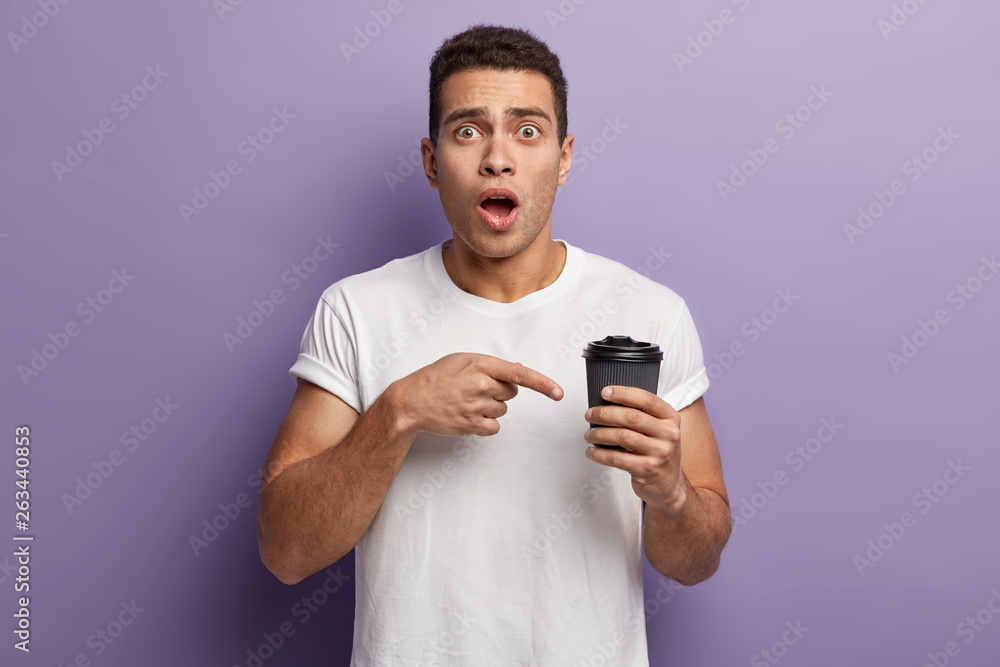 Shot of stupefied young guy points finger at takeaway coffee, surprised to have empty paper cup without any drink, wears white casualt shirt, has opened mouth, isolated against purple background. Shot of stupefied young guy points finger at takeaway coffee, surprised to have empty paper cup without any drink, wears white casualt shirt, has opened mouth, isolated against purple background.