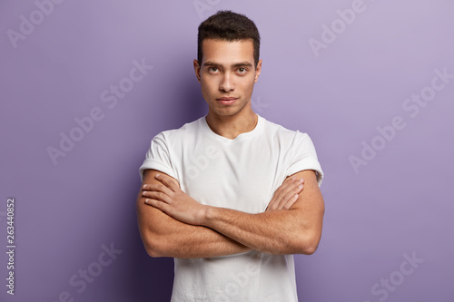 Photography Self assertive serious young guy keeps muscular arms crossed over chest, looks directly at camera, gazes attentively something, dressed in casual white t shirt, looks confident