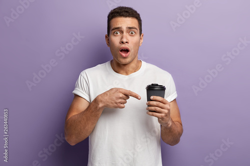 Shot of stupefied young guy points finger at takeaway coffee, surprised to have empty paper cup without any drink, wears white casualt shirt, has opened mouth, isolated against purple background. © wayhome.studio 