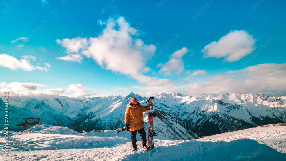 A young guy in a yellow down jacket with a snowboard in his hands stands on the top of the mountain. Snowboarder stands on the background of a beautiful view of the snow-capped mountains. Snowboarder 