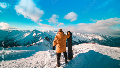 A young guy in a yellow down jacket with a snowboard in his hands stands on the top of the mountain. Snowboarder stands on the background of a beautiful view of the snow-capped mountains. Snowboarder 