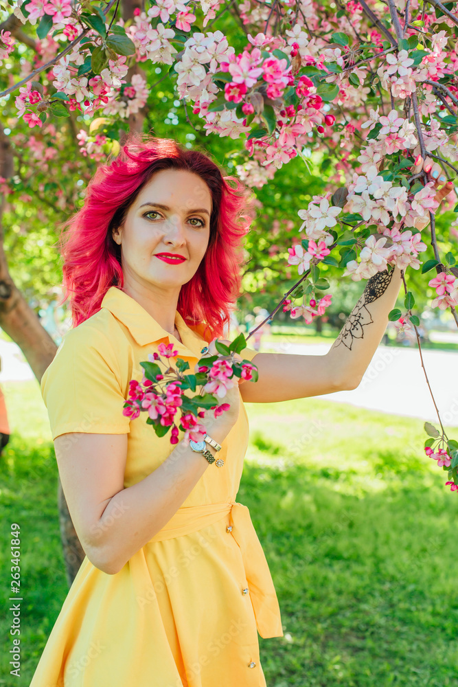 Beautiful and young woman with bright red hair and ref lips standing next to a blooming apple tree in a yellow dress.
