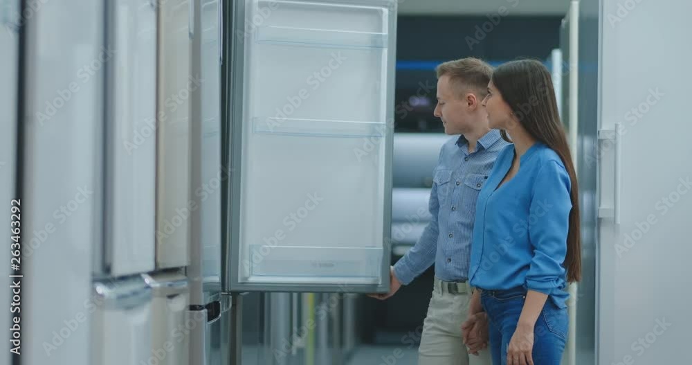 A young married couple open the door of the new refrigerator inspect the design and quality before buying in a consumer electronics store. Read the information on the price tag and characteristics A young married couple open the door of the new refrigerator inspect the design and quality before buying in a consumer electronics store. Read the information on the price tag and characteristics