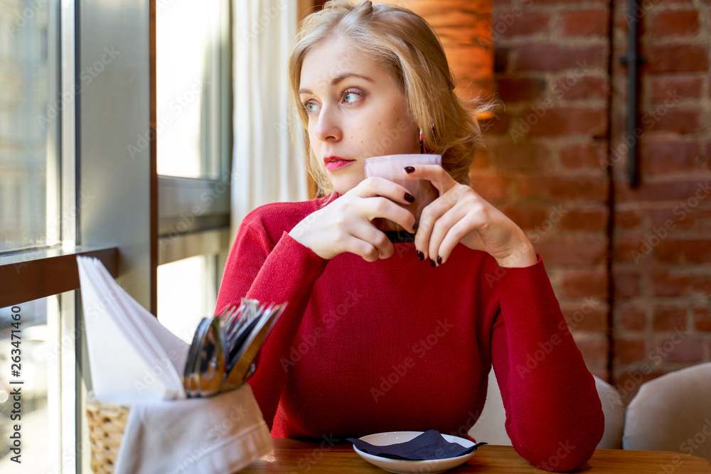 Beautiful blonde girl looking out the window, holding a Cup of coffee. Pensive woman thinking about upcoming cases Beautiful blonde girl looking out the window, holding a Cup of coffee. Pensive woman thinking about upcoming cases