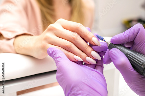 Manicurist work on a woman client hands, make her nails look beautiful. Salon procedure in process. Professional works in gloves using drill for sterility